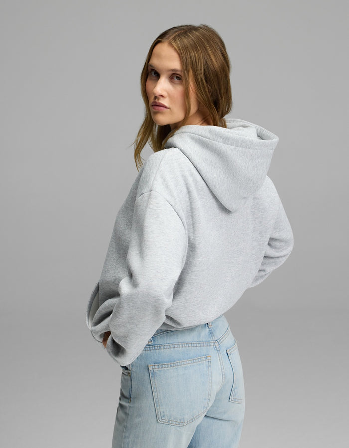 A woman with light brown hair models the Cozy Earth Women’s CityScape Hoodie in light gray, paired with light blue jeans. She stands turned slightly to the side, glancing back over her shoulder against a plain gray background.|Color:Heather Grey