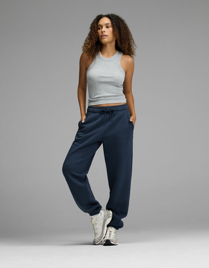 A woman with curly hair wears a gray sleeveless top, Cozy Earth’s Women’s CityScape Sweatpant in navy blue, and white sneakers. She stands with one foot slightly raised and her hands in her pockets against a plain gray background.|Color:Eclipse