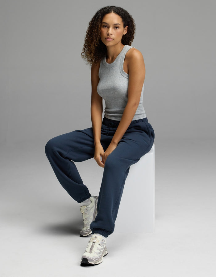 A person with curly hair, wearing a gray tank top, Cozy Earth Women’s CityScape Sweatpant in navy, and sneakers, sits on a white block against a plain gray background, looking at the camera.|Color:Eclipse