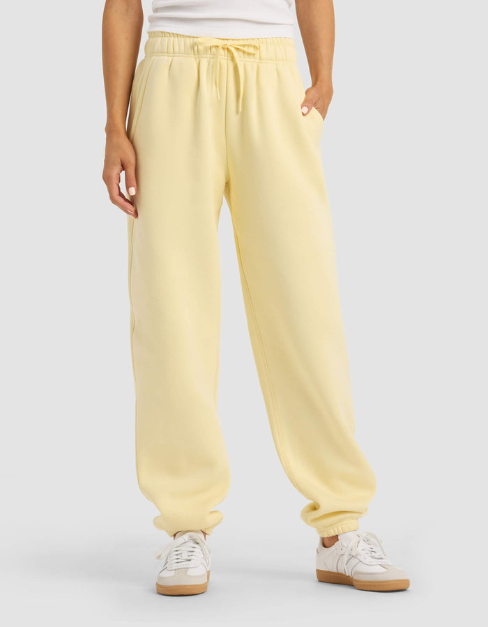 A person stands against a plain background with hands in the pockets of Cozy Earth Women's Cityscape Sweatpant in light yellow, paired with a white top and white sneakers.|Color:Meringue