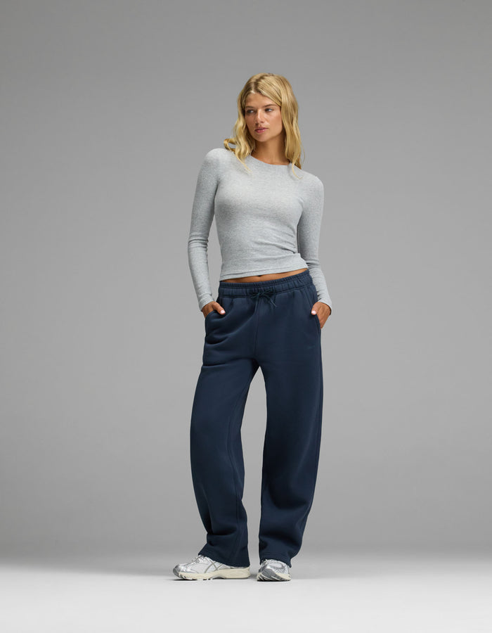 A woman with blonde hair wearing a light gray long-sleeve shirt, Cozy Earth Women’s Cityscape Wide Leg Pants in navy blue, and white sneakers stands with her hands in her pockets against a plain gray background.|Color:Eclipse