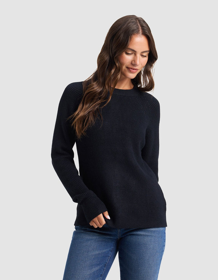 A woman with long brown hair models the Cozy Earth Women's Classic Crewneck, wearing blue jeans and standing against a light gray background, looking down with a slight smile and one hand in her pocket. |Color:Jet Black