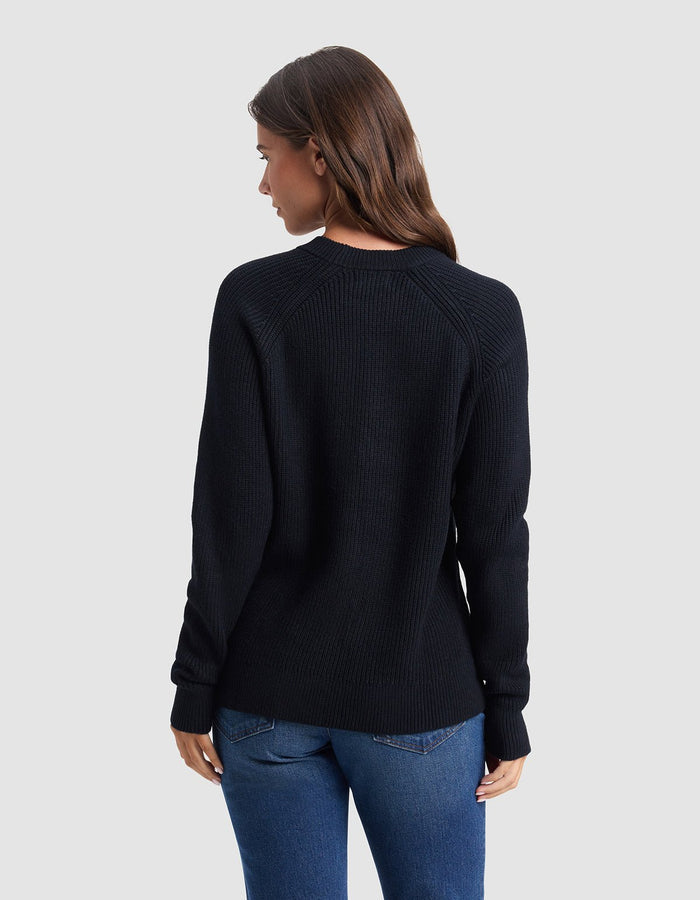 A woman with long brown hair, wearing the Cozy Earth Women's Classic Crewneck and blue jeans, stands facing away from the camera against a plain light gray background.|Color:Jet Black