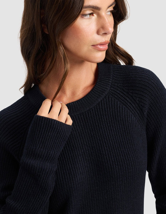 A woman with wavy brown hair wears the Cozy Earth Women's Classic Crewneck in dark navy, facing slightly to the side with her hand near her collar. The background is light gray.|Color:Jet Black