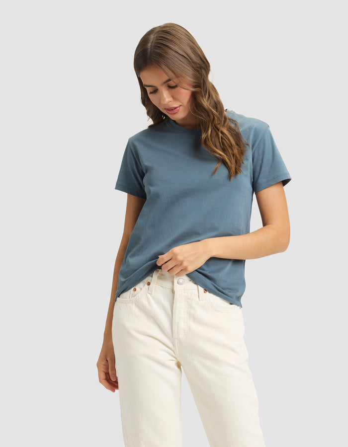 A woman with wavy brown hair wears the Cozy Earth Women's Easy Tee in blue and off-white jeans, gently holding the shirt's hem against a light gray background. |Color:Dawn