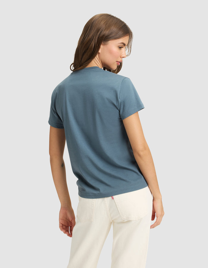A woman with light brown hair models the Cozy Earth Women's Easy Tee in blue and off-white pants, standing with her back partially turned against a plain light gray background. |Color:Dawn