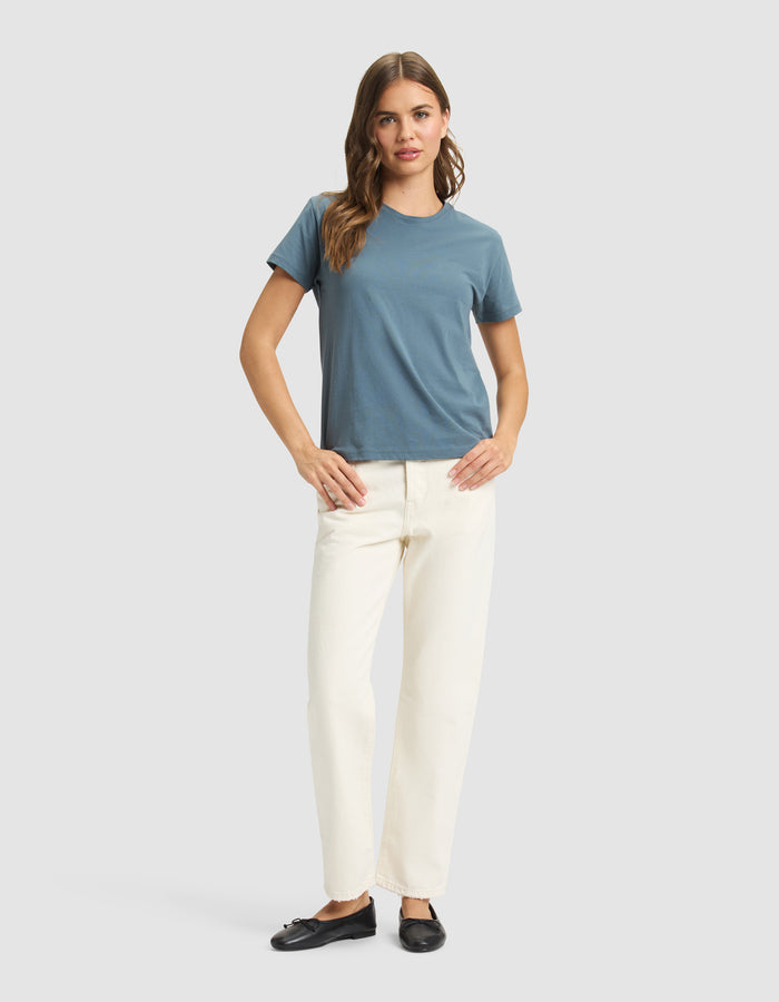 A woman with long, wavy hair stands facing forward against a plain light gray background, wearing the Cozy Earth Women's Easy Tee in blue, off-white pants, and black slip-on shoes, with her hands resting on her hips. |Color:Dawn
