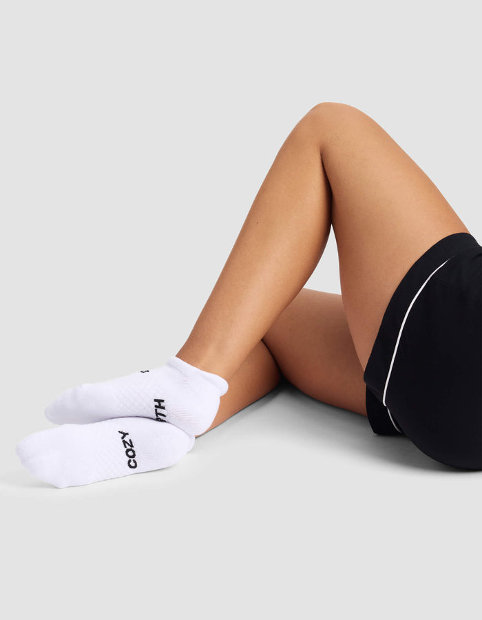 A woman wears Cozy Earth Women's Essential Ankle Socks with black text, paired with black shorts, sitting cross-legged on the floor against a light grey background.|Color:White