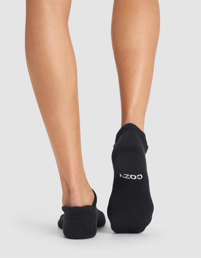A person stands barefoot on a white background, wearing black Essential Ankle Socks by Cozy Earth. The back of their legs and feet are visible, with "COZY" printed in white on the sole of one sock. |Color:Black