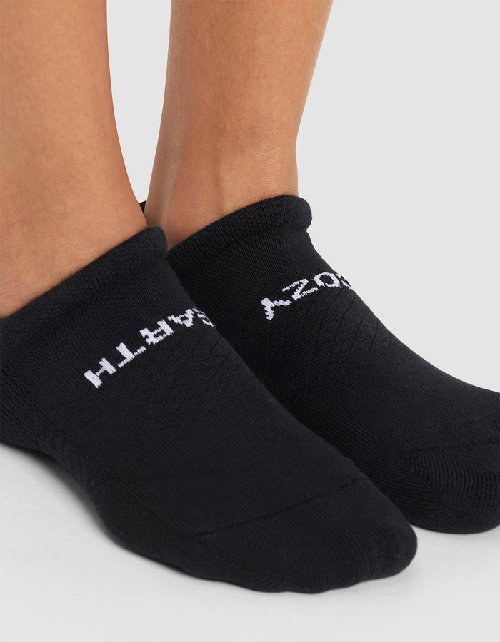 A close-up of two feet wearing Cozy Earth Essential Ankle Socks, black with white text on top, standing on a light gray surface. The sock text is partially visible. |Color:Black