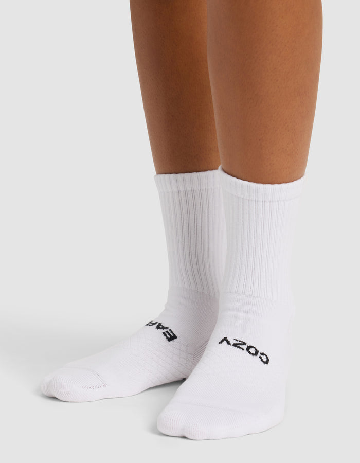 Someone wearing Cozy Earth Essential Calf Socks stands on a light surface. The white ribbed crew socks feature partially visible dark text on top, with only their lower legs and feet shown.|Color:White