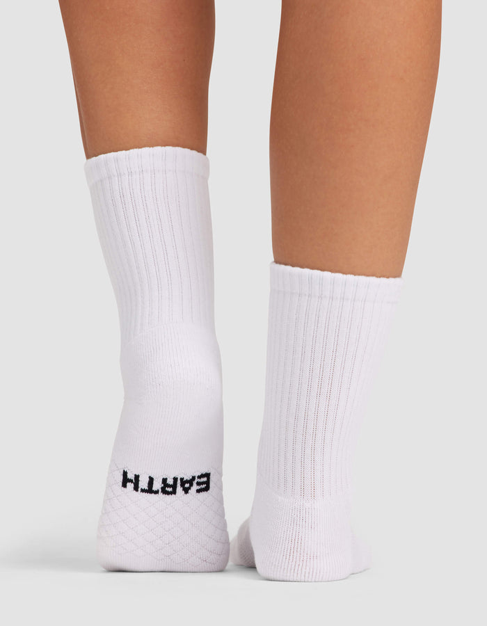 A close-up shows the back of two legs wearing Cozy Earth Women's Essential Calf Socks in white ribbed fabric. The left sock features "EARTH" in dark stitching on the heel, set against a plain light gray background.|Color:White