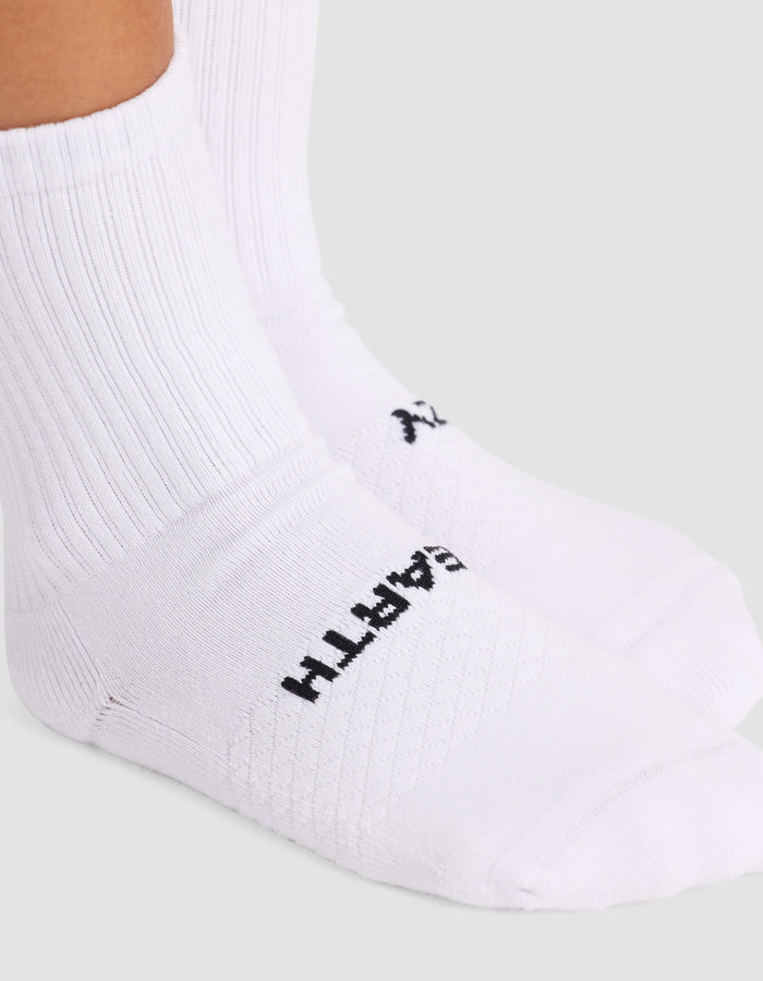 A close-up shows a person wearing Cozy Earth Women’s Essential Calf Socks in white with ribbing and a logo near the toes. The background is plain and light-colored.|Color:White
