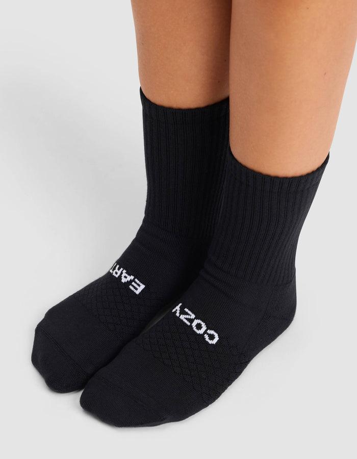 A person wearing Cozy Earth's Women's Essential Calf Sock in black, featuring "COZY EARTH" text on the tops of the feet, stands on a light gray surface.|Color:Black