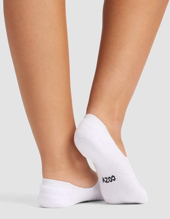 Close-up of a person’s lower legs and feet wearing Cozy Earth Women's Essential No-Show Socks with "COZY" in black on the sole, standing against a plain light gray background.|Color:White