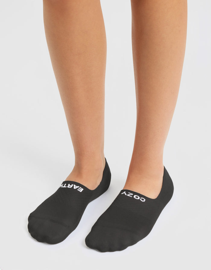 A person wears Cozy Earth's Essential No-Show Sock 4-Pack in black, featuring "EAZY" and "COZY" printed in white on top, standing against a white background.
