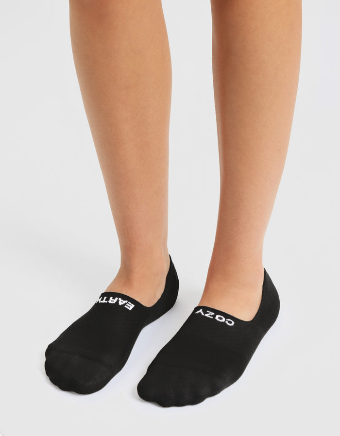 A person stands on a white surface wearing Cozy Earth Women's Essential No-Show Socks in black, with "EARTH" in white on the left sock and "COZY" in white on the right.|Color:Black