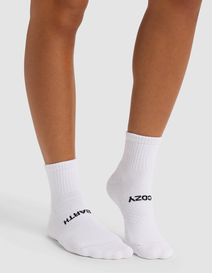 A person stands wearing Cozy Earth Essential Quarter Socks in white. "COZY" is written in black on the top of one sock and "EARTH" on the other. The background is plain and light-colored.|Color:White