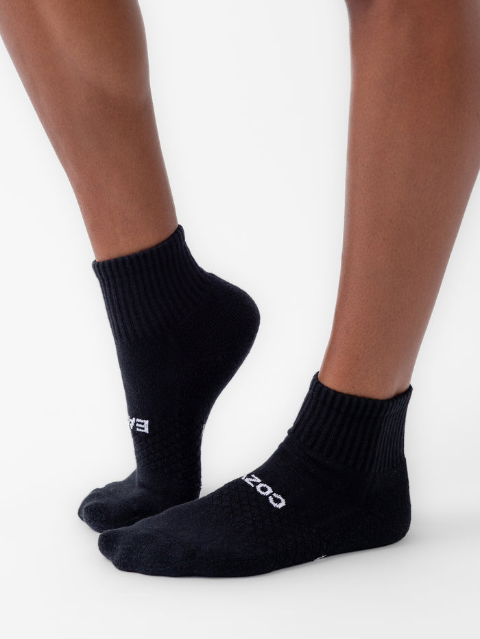 A person wearing black, quarter-length socks stands against a white background. The socks, from the Essential Quarter Sock 2-Pack by Cozy Earth, feature a ribbed top and textured pattern with the brand name "COZY" visible on the side. The person's skin is light brown. |Color:Black