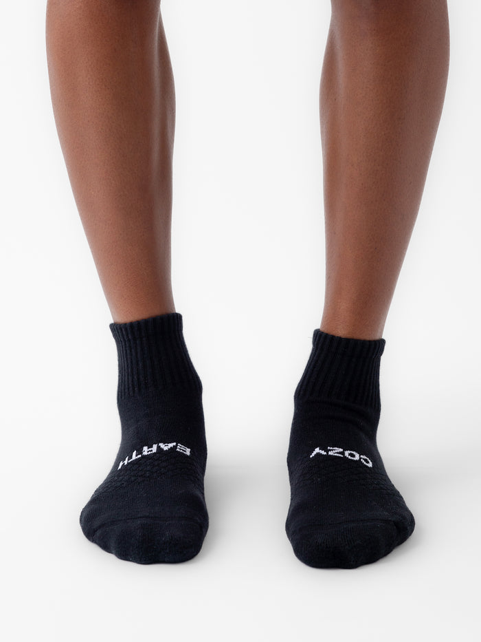 A person wears black Cozy Earth Essential Quarter Socks on a white surface. The socks have white text: "EARTH" on the left toe and "COZY" on the right. |Color:Black