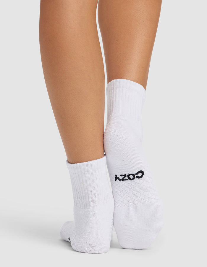 A person stands with feet crossed, wearing Cozy Earth Essential Quarter Socks in white. "COZY" is written in black on the sole of one sock. The background is plain and light-colored.|Color:White