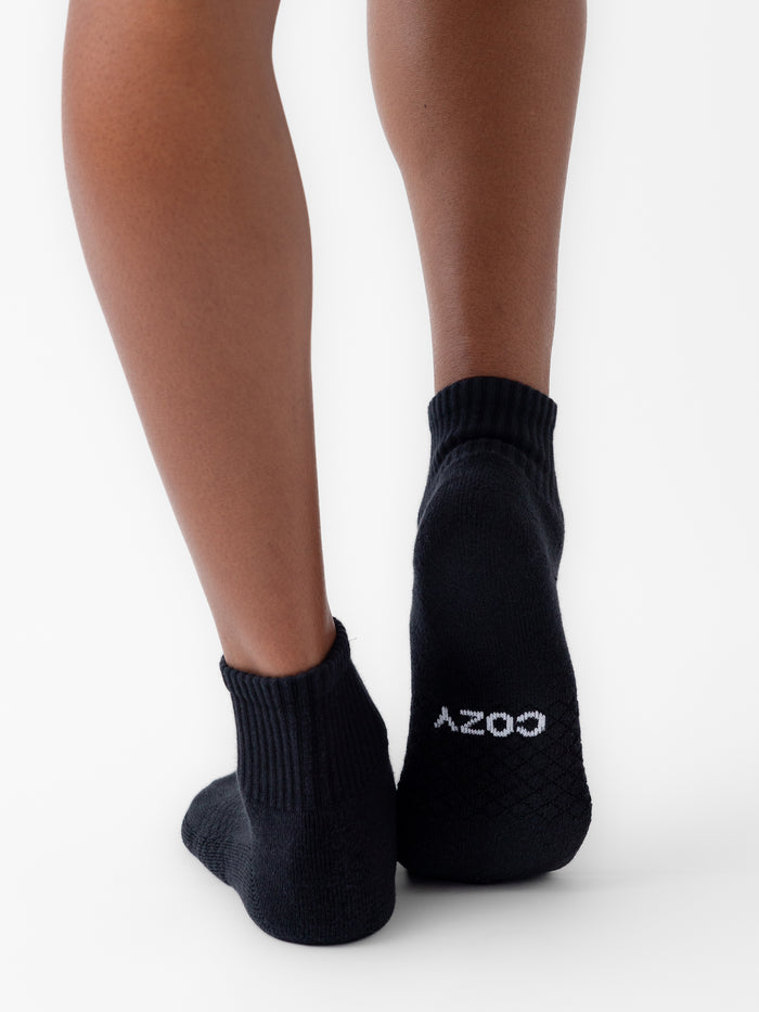 A person wearing Cozy Earth’s Essential Quarter Sock (black, from the 8-pack) stands on a white background. Only the lower legs and feet are shown, with "COZY" in white text visible on the sole of one sock. |Color:Black