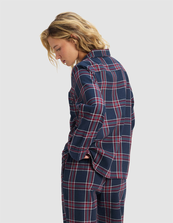 A woman with blond hair stands in profile with hands in pockets, wearing the Cozy Earth Women's Farmhouse Long Sleeve Pajama Top in navy blue and red plaid, paired with matching pants against a plain light gray background.|Color:Eclipse Plaid