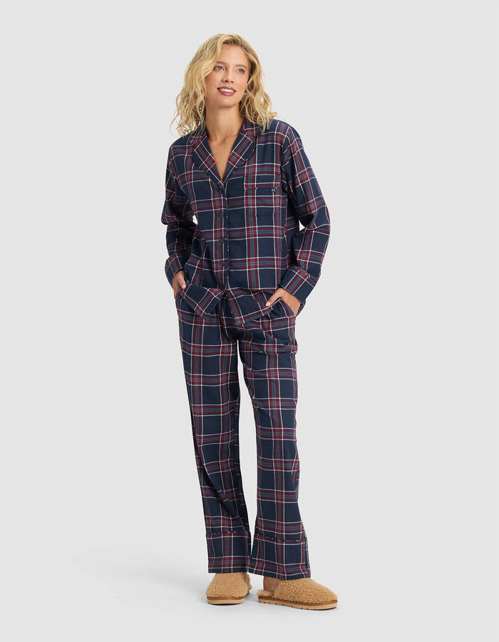 A person stands smiling, wearing the Cozy Earth Women's Farmhouse Long Sleeve Pajama Top in navy blue with red and white plaid, paired with loose pajama pants and tan fuzzy slippers. The top features buttons and pockets.|Color:Eclipse Plaid