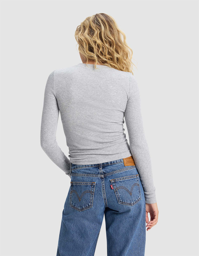 A woman with wavy blonde hair, wearing Cozy Earth's Women's Fine Ribbed Long Sleeve Tee in light gray and blue jeans, stands with her back to the camera against a plain light background.|Color:Heather Grey