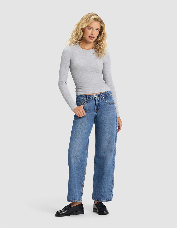 A woman with long blonde curly hair wears the Cozy Earth Women's Fine Ribbed Long Sleeve Tee, styled with blue high-waisted wide-leg jeans and black loafers, standing against a plain light background.|Color:Heather Grey