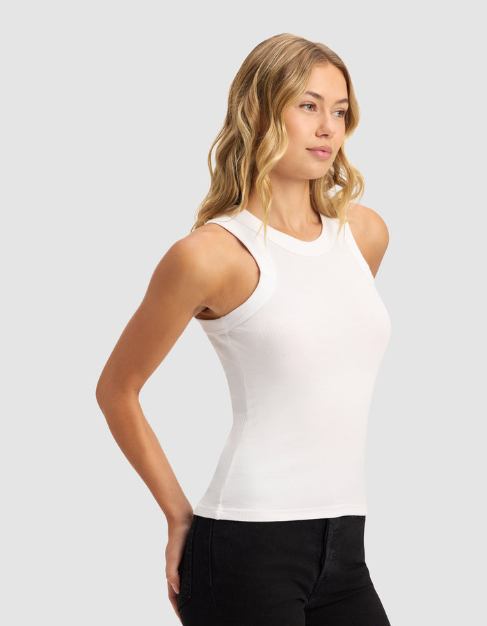 A woman with wavy blonde hair wears the Cozy Earth Women's Fine Ribbed Tank in white and black pants, standing against a plain light gray background, looking slightly to the side with her hands behind her back.|Color:White