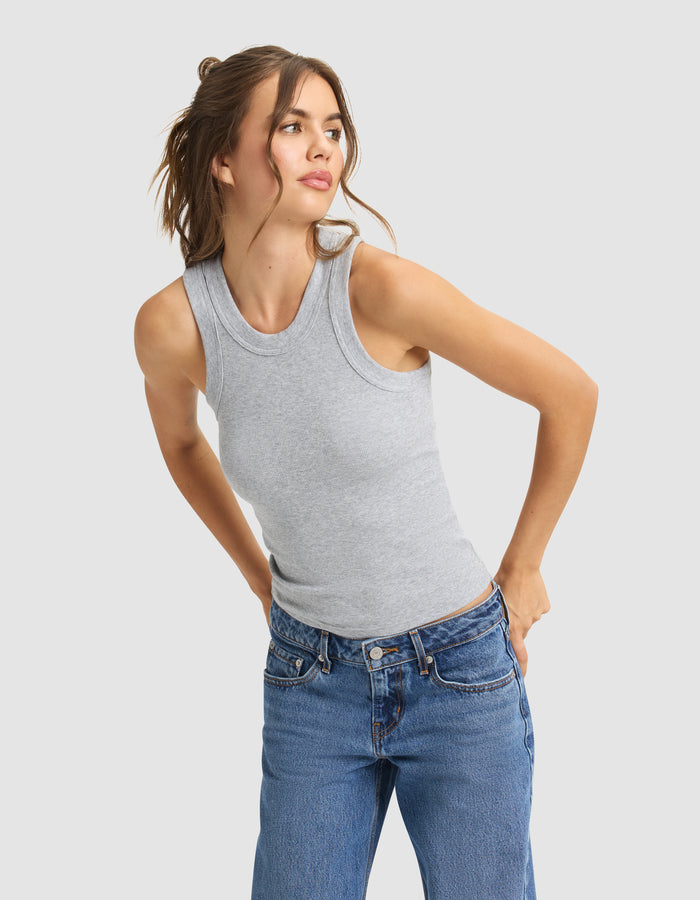 A woman with brown hair in a loose ponytail wears Cozy Earth's Women's Fine Ribbed Tank in gray and blue jeans, standing against a light background and looking slightly left with her hands on her hips.|Color:Heather Grey