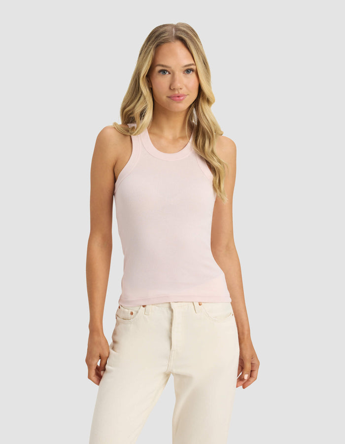 A woman with long blonde hair wears Cozy Earth's Women's Fine Ribbed Tank in light pink and off-white jeans, standing against a plain light gray background and facing forward with a neutral expression.|Color:Ballet