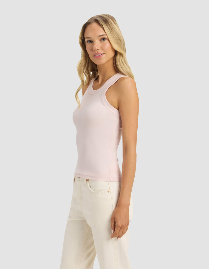 A woman with long blonde hair wears the Cozy Earth Women's Fine Ribbed Tank in light pink with off-white pants, standing against a plain light gray background and smiling softly.|Color:Ballet