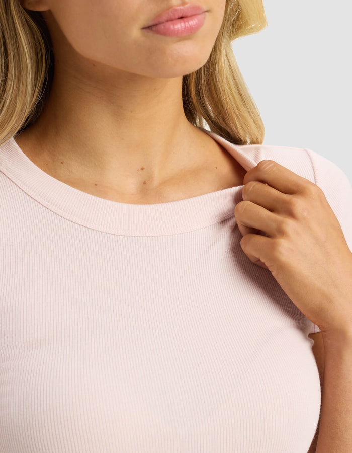 A woman wears the Cozy Earth Women's Fine Ribbed Tee in light pink, gently pulling at the collar with her right hand. Only her lower face, neck, and upper torso are visible against a plain light background.|Color:Ballet