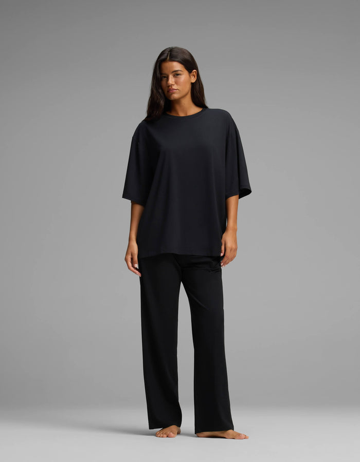 A woman stands barefoot against a gray background, wearing the Cozy Earth Women’s First Layer Boyfriend Sleep Tee & Pajama Pant Set in black, with her hair down and her arms relaxed by her sides.|Color:Jet Black