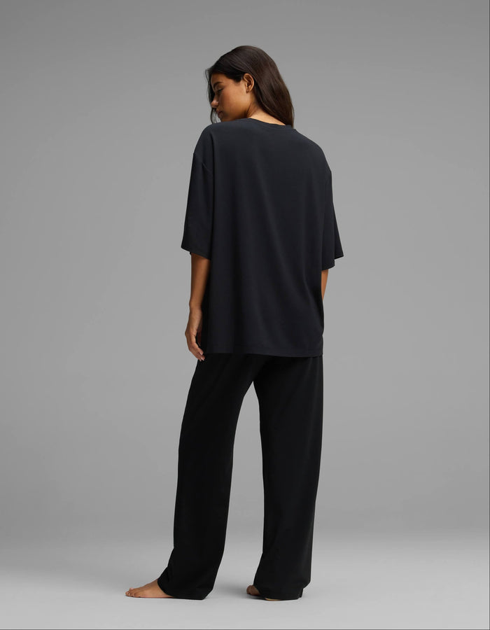 A woman stands barefoot, facing away, wearing the Cozy Earth Women’s First Layer Boyfriend Sleep Tee & Pajama Pant Set in black against a plain gray background.|Color:Jet Black