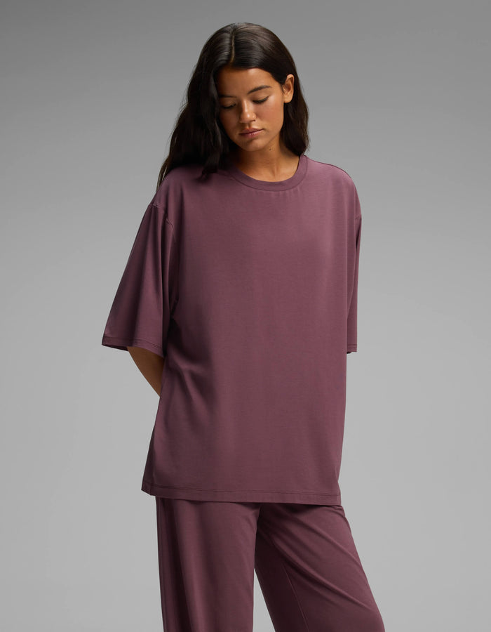 A woman with long dark hair stands against a plain gray background, her hands behind her back, wearing the Cozy Earth Women’s First Layer Boyfriend Sleep Tee & Pajama Pant Set in a loose mauve fit.|Color:Fig