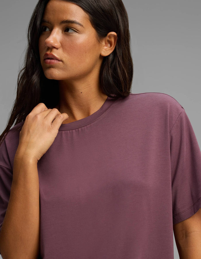 A woman with long dark hair wearing the Cozy Earth Women’s First Layer Pajama Short Sleeve Tee in mauve looks to the side against a plain gray background.|Color:Fig