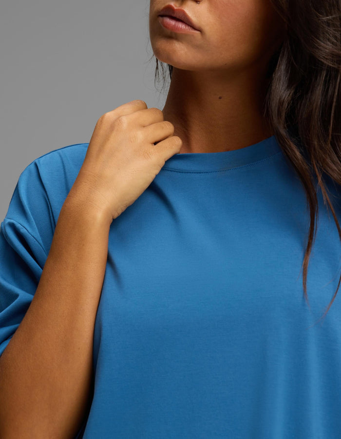 A person wearing the Cozy Earth Women’s First Layer Boyfriend Sleep Tee in blue touches the collar with one hand. The lower part of their face and dark hair are visible against a plain, neutral background.|Color:Anchor