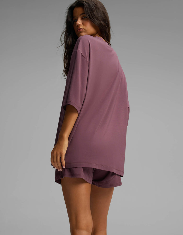 A woman with long hair stands sideways and looks over her shoulder against a plain gray background, wearing the Cozy Earth Women’s First Layer Boyfriend Sleep Tee in mauve.|Color:Fig