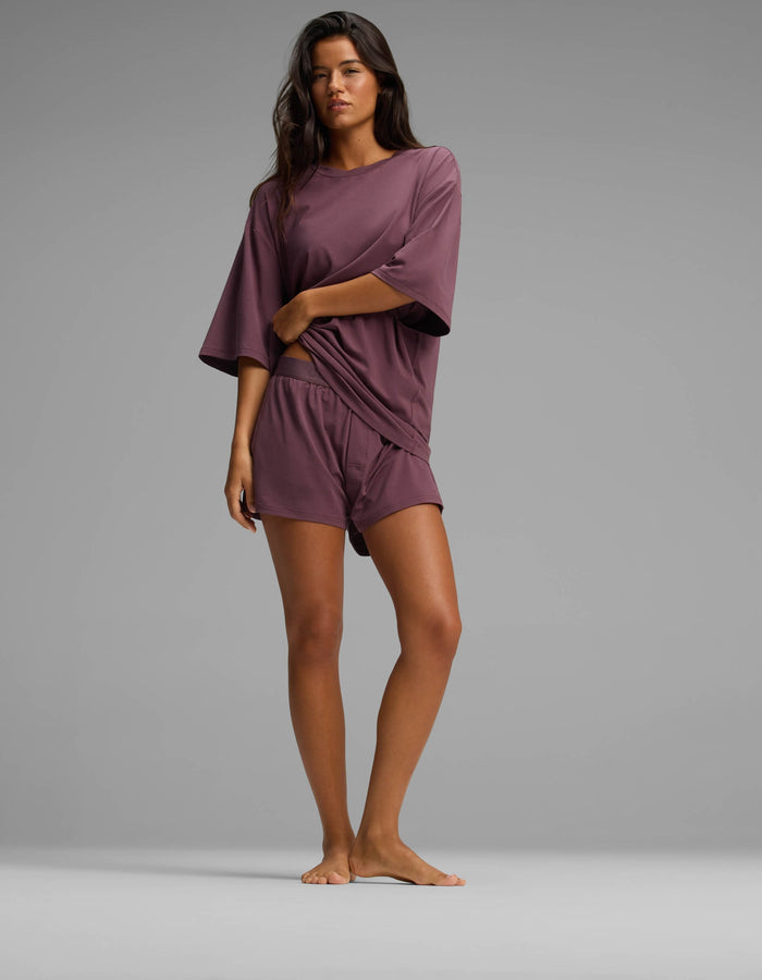 A woman stands barefoot on a plain background, wearing the Cozy Earth Women’s First Layer Boyfriend Sleep Tee in mauve with matching shorts, holding the hem of her shirt with one hand.|Color:Fig