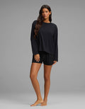 A woman with long brown hair stands barefoot on a grey background, modeling the Cozy Earth Women’s First Layer Long Sleeve Sleep Tee & Boxer Short Set in black, looking at the camera with a neutral expression.|Color:Jet Black