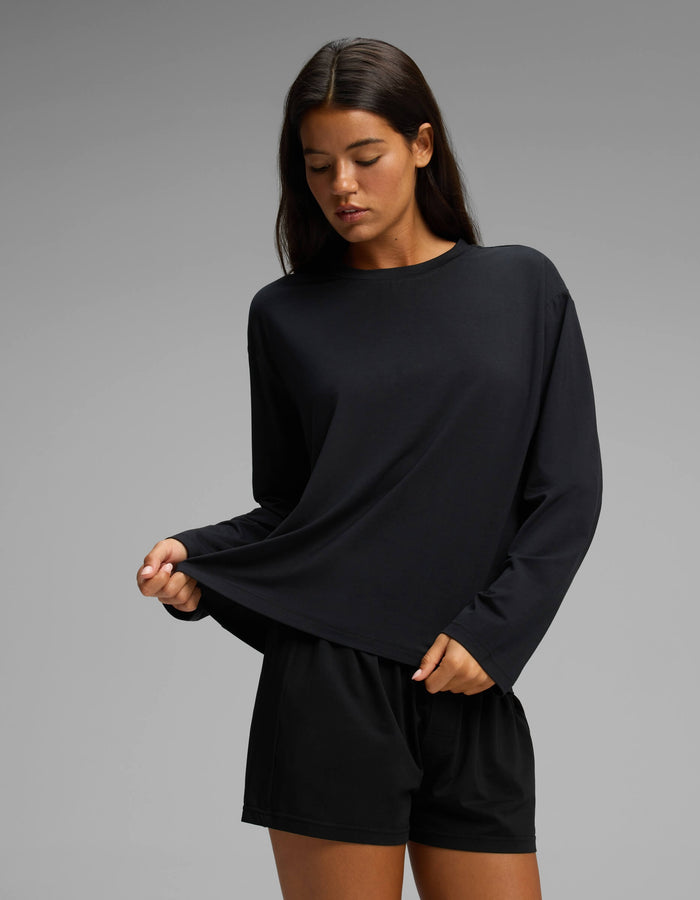 A woman stands against a plain gray background, gently pulling at the hem of her black Cozy Earth Women’s First Layer Long Sleeve Sleep Tee & Boxer Short Set while looking down.|Color:Jet Black