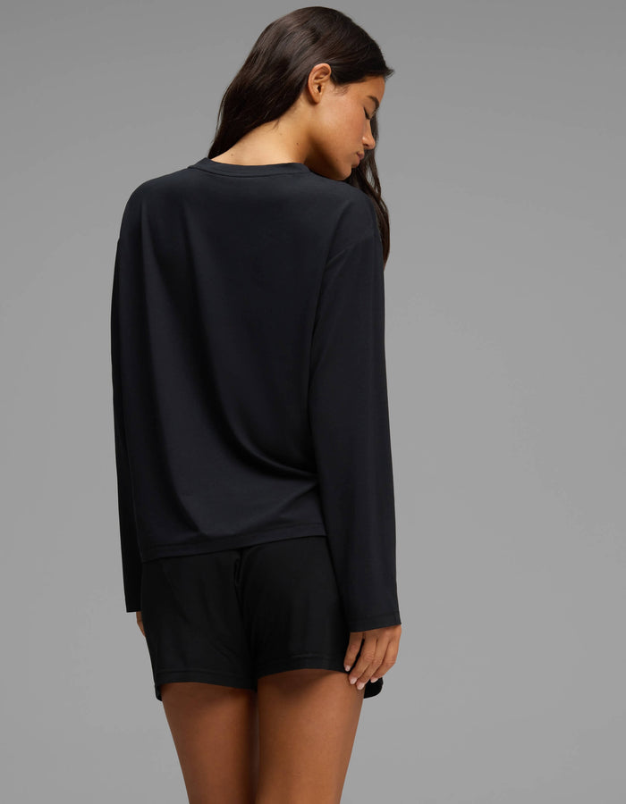 A woman with long brown hair is seen from behind, looking down, wearing the Cozy Earth Women’s First Layer Long Sleeve Sleep Tee & Boxer Short Set in black against a plain gray background.|Color:Jet Black