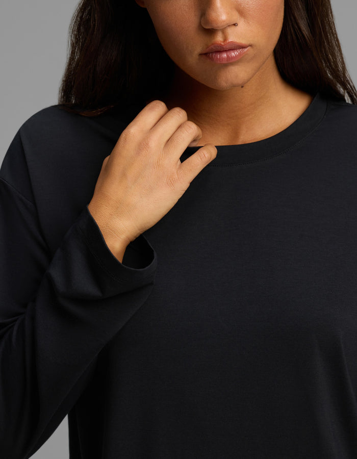 A woman wearing the Cozy Earth Women’s First Layer Long Sleeve Sleep Tee in black touches her collar with her right hand. Only the lower half of her face, neck, and upper torso are visible against a neutral gray background.|Color:Jet Black