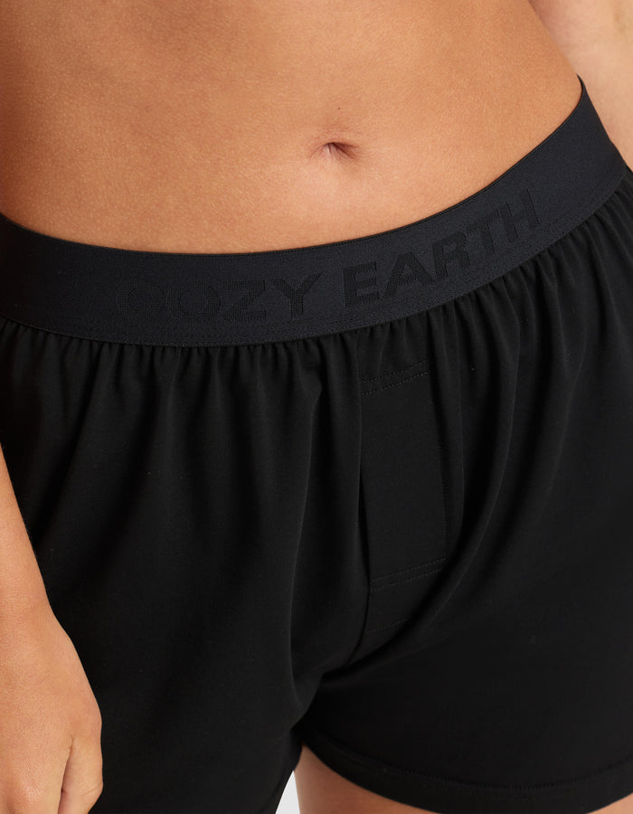 Close-up of a person wearing Cozy Earth Women's First Layer Pajama Short Sleeve Tee & Boxer Short Set, featuring black boxer shorts with a waistband embossed with “COZY EARTH.” Only the lower torso and upper thighs are visible.|Color:Jet Black