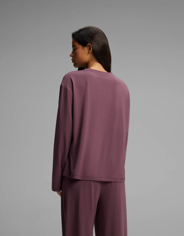 A woman with long dark hair, wearing the Cozy Earth Women’s First Layer Long Sleeve Sleep Tee and matching pants in mauve, stands facing away from the camera against a plain gray background.|Color:Fig