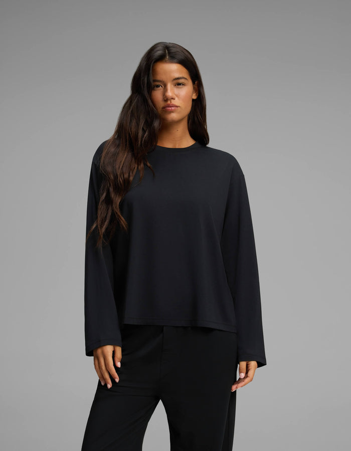 A woman with long dark hair wears the Cozy Earth Women’s First Layer Long Sleeve Sleep Tee in black, paired with black pants, standing against a plain gray background.|Color:Jet Black