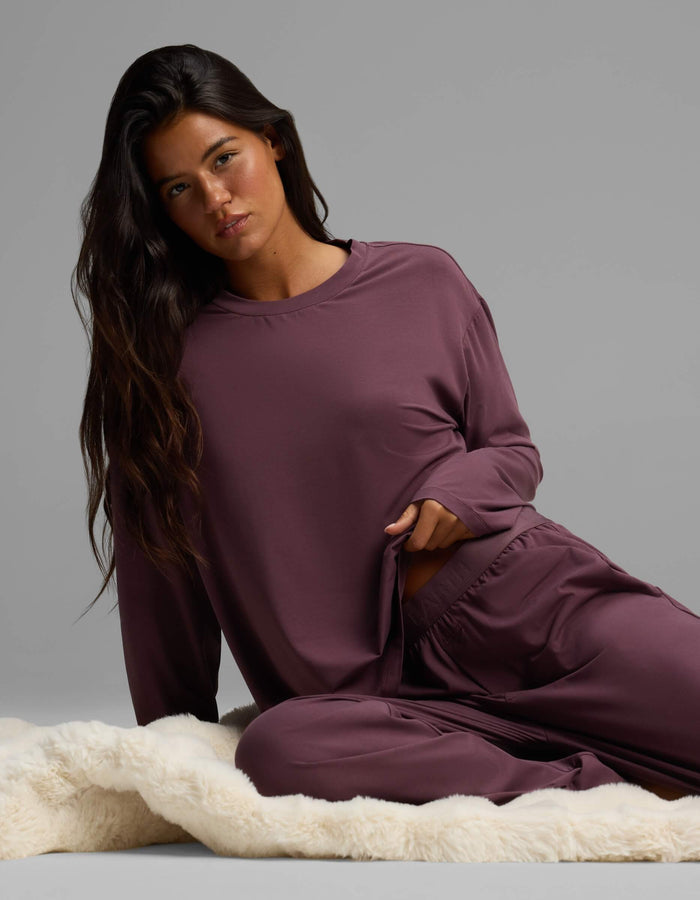 A woman with long dark hair sits on a cream-colored rug, wearing the Cozy Earth Women’s First Layer Long Sleeve Sleep Tee in mauve with matching pants, looking at the camera with a neutral expression.|Color:Fig